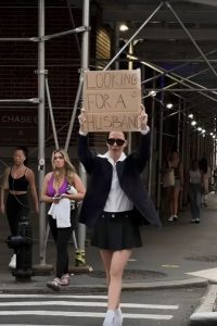 A woman is walking down the street with a large sign that says, “looking for a husband,” when something unexpected occurs.